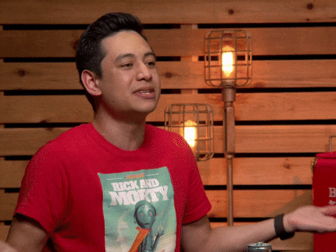 Man in a red Rick and Morty T-shirt talks and gestures with an open hand in front of a wooden wall with caged lamps